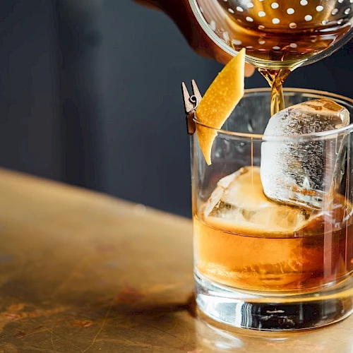 A whiskey glass with large ice cubes, a citrus peel garnish, and a pour from a bottle into a bar setting.