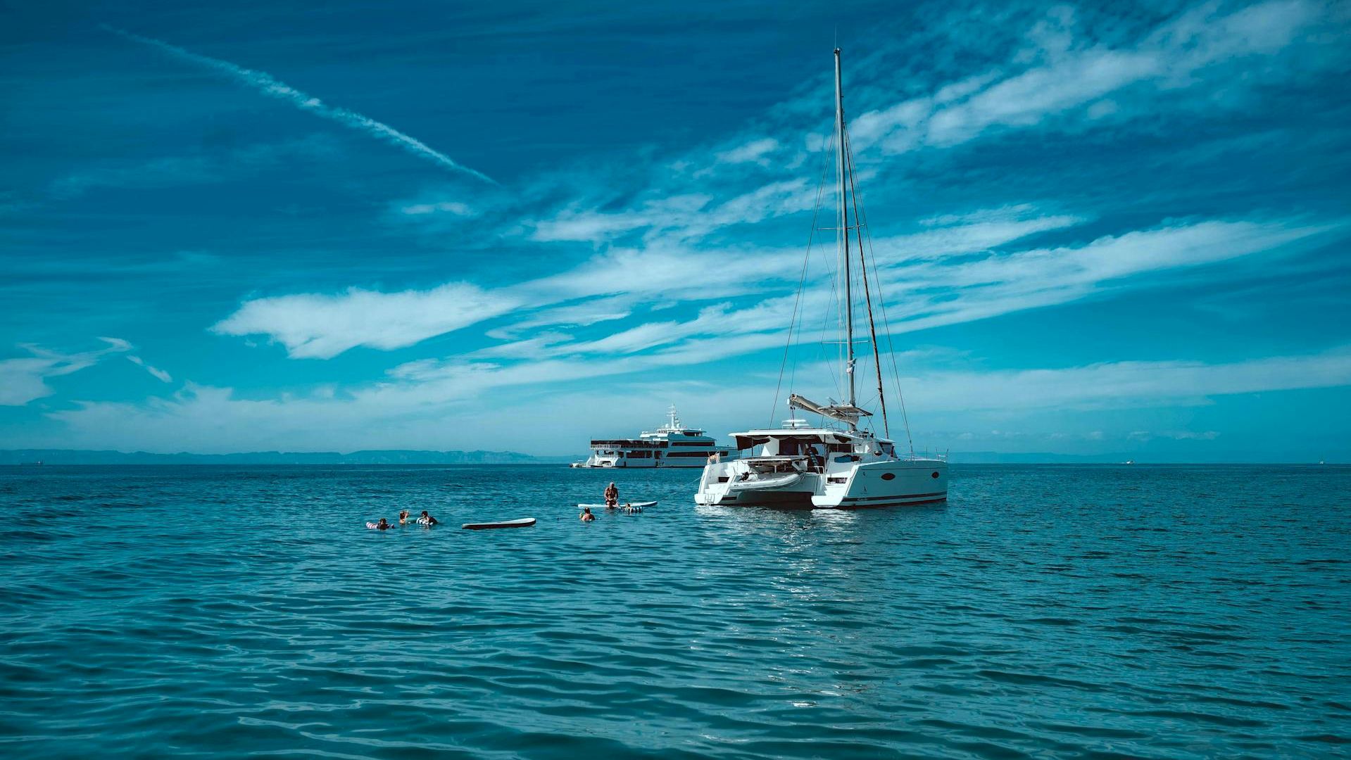 A sunny sea scene with a white sailboat anchored near a smaller yacht, calm turquoise water, and a blue sky with wispy clouds.