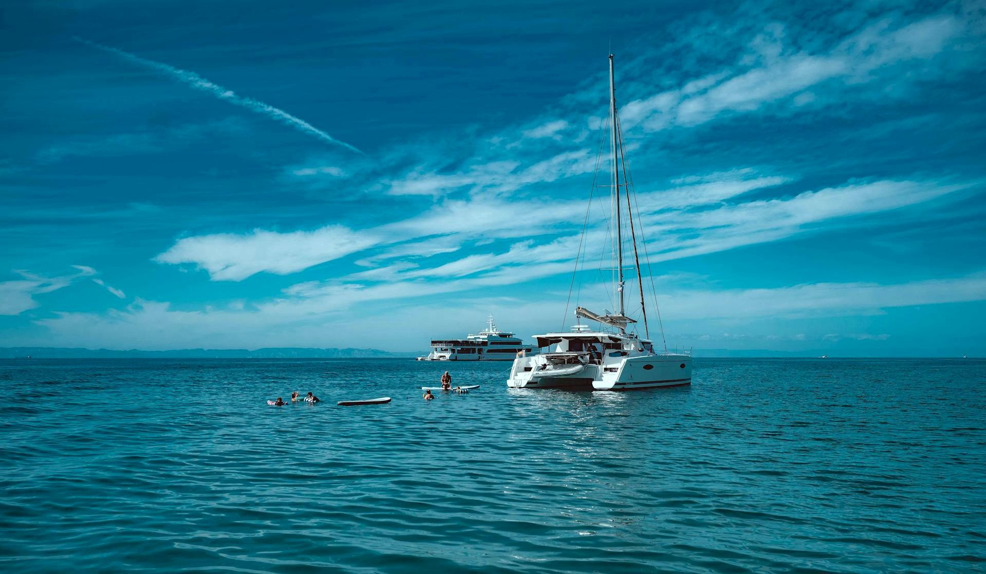 A sunny sea scene with a white sailboat anchored near a smaller yacht, calm turquoise water, and a blue sky with wispy clouds.