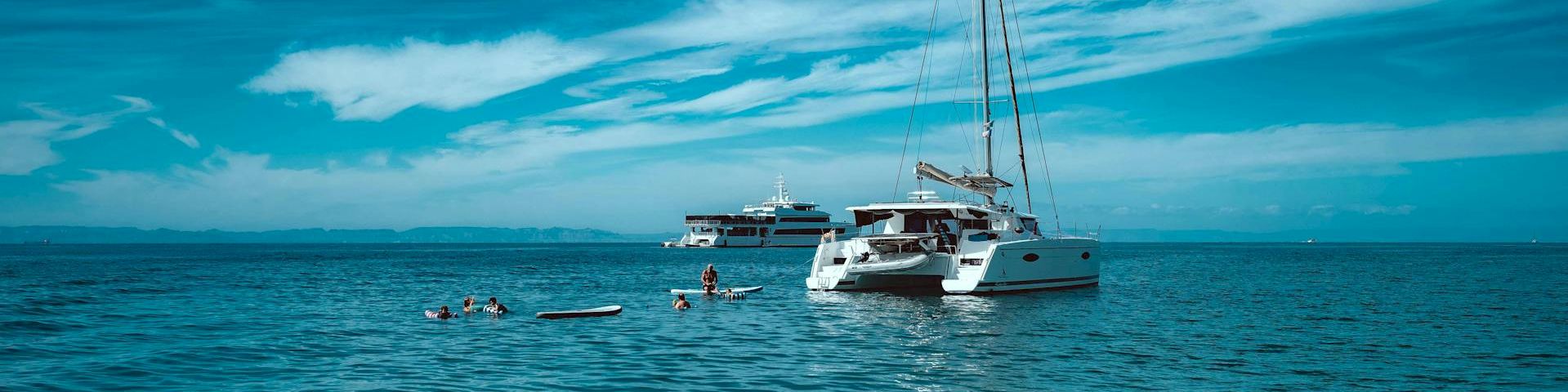 A sunny sea scene with a white sailboat anchored near a smaller yacht, calm turquoise water, and a blue sky with wispy clouds.