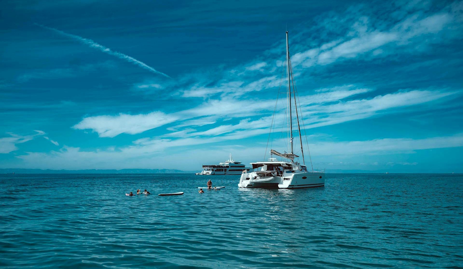 A calm sea with two sailing cats and a larger white yacht anchored near the horizon under a blue sky with scattered clouds, swimmers nearby.