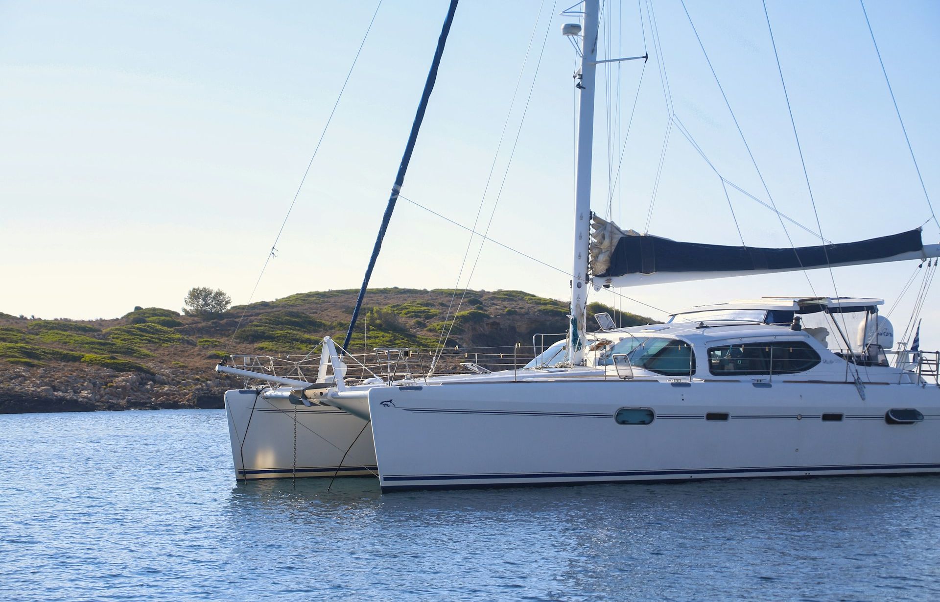 Two white sailboats anchored together in calm blue water near a rocky shoreline under a clear sky.