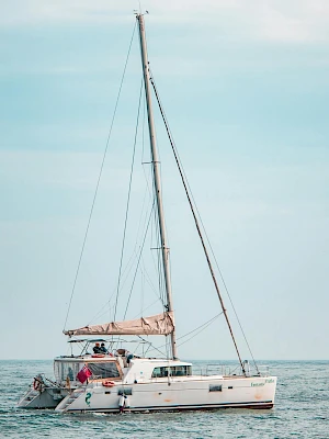 A white catamaran/sailboat steers calmly on blue ocean water under a clear sky, with a few people on deck enjoying a sunny day, trailing sail lines.