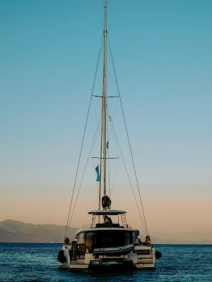 A sailboat anchored on calm blue water at sunset, rear view with people on deck and a tall mast against a clear sky.