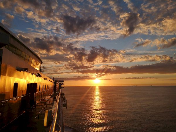 Sunset over calm waters from a ship&rsquo;s side, warm orange hues glaze the deck as clouds catch fire and the horizon sinks into golden serenity.
