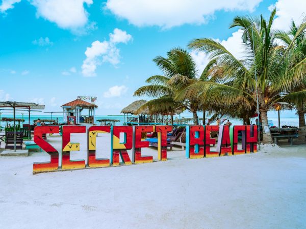 Colorful &ldquo;SECRET BEACH&rdquo; sign on white sand, palm trees, and turquoise sea under a bright blue sky with a few clouds.