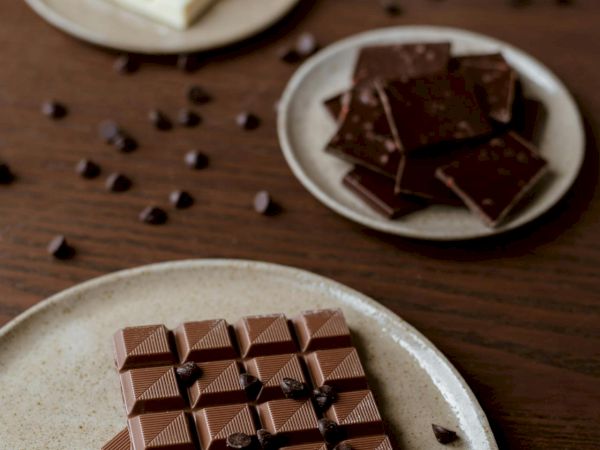 Assorted chocolate: white, milk, and dark bars cut into squares, scattered cacao nibs on a wooden table, with a small bowl of extra bits.