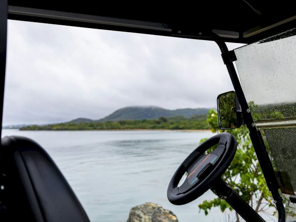 A boat cockpit view looking out over calm water with distant hills, steering wheel on the right, wet window, and rocky shore nearby.