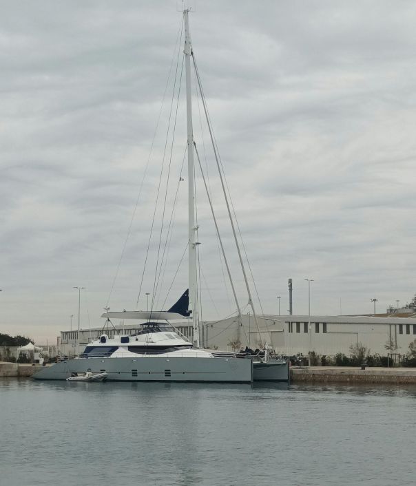 A sleek white sailboat with a tall mast docked by a calm harbor, other small boats nearby and a low-lying industrial/port backdrop.