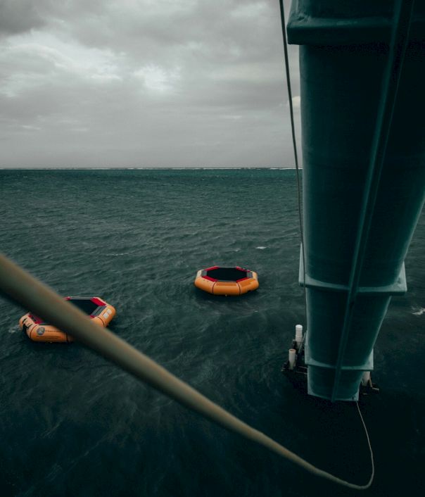A high-angle view over the sea shows a pier or mast with a rope, and two orange life buoys floating in the dark, choppy water below.
