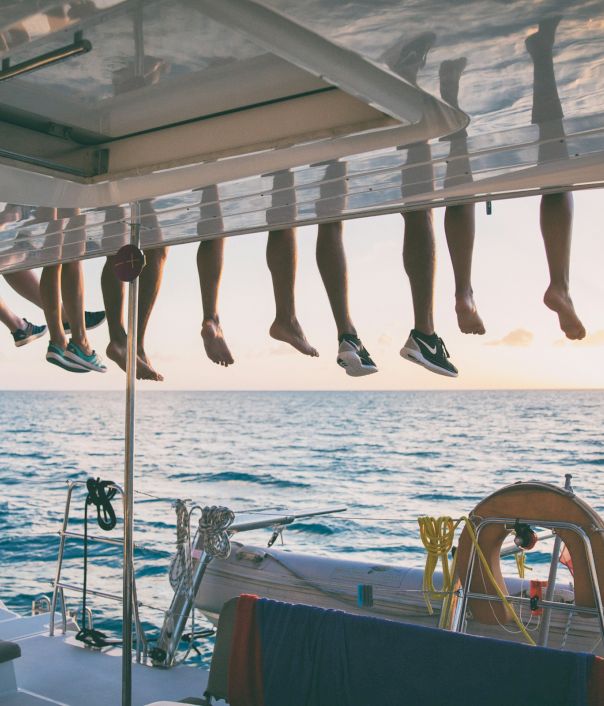 People&rsquo;s legs hang over the edge of a boat at sunset, while the rest of the boat and ocean stretch out below.