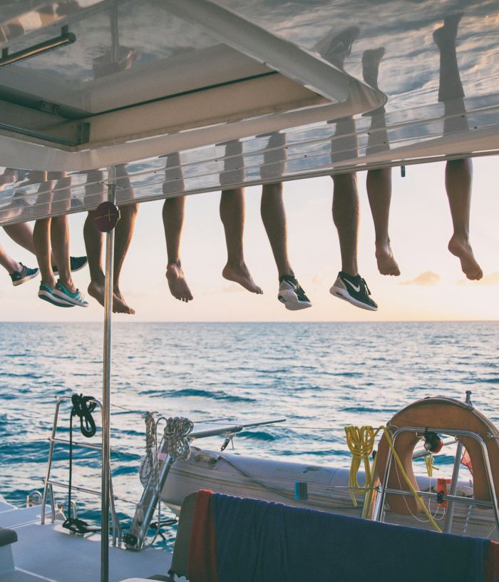People&rsquo;s legs hang over the edge of a boat at sunset, while the rest of the boat and ocean stretch out below.