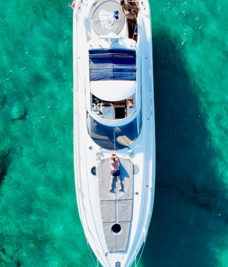 A white speedboat glides over clear turquoise water, sunbathers lounging on deck, a parasol and blue cushions catching the light.