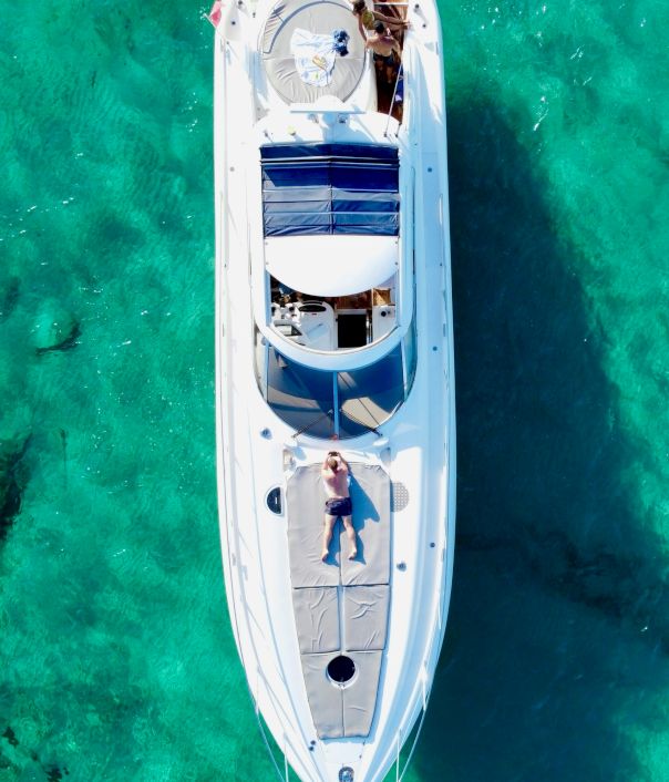 A white speedboat glides over clear turquoise water, sunbathers lounging on deck, a parasol and blue cushions catching the light.
