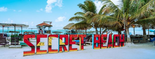 A tropical beach with white sand, turquoise water, palm trees, and a colorful sign spelling &ldquo;SOUTH&rdquo; on the shore.