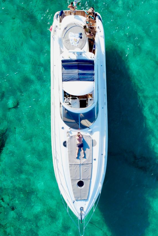 A white luxury yacht anchored in clear turquoise water, viewed from above, with people relaxing on deck. End of sentence.