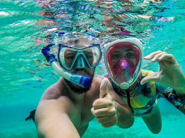 Two people snorkeling underwater, wearing masks and snorkels, giving a thumbs up and a close-up selfie in clear turquoise water.