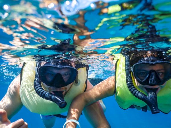Two people snorkeling underwater with masks and snorkels, wearing life vests, reaching toward the camera in clear blue water.