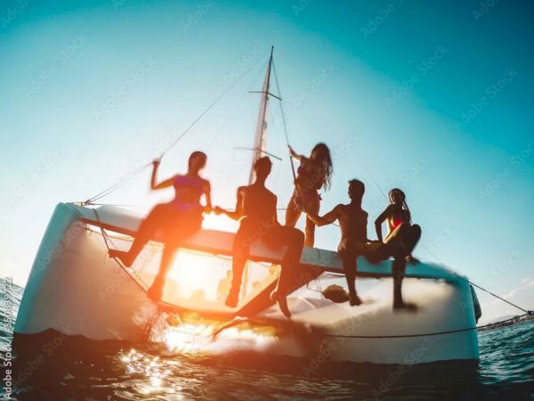 A group of people sitting on a sunlit sailboat tilted on the water, silhouettes against a bright, blue sky.
