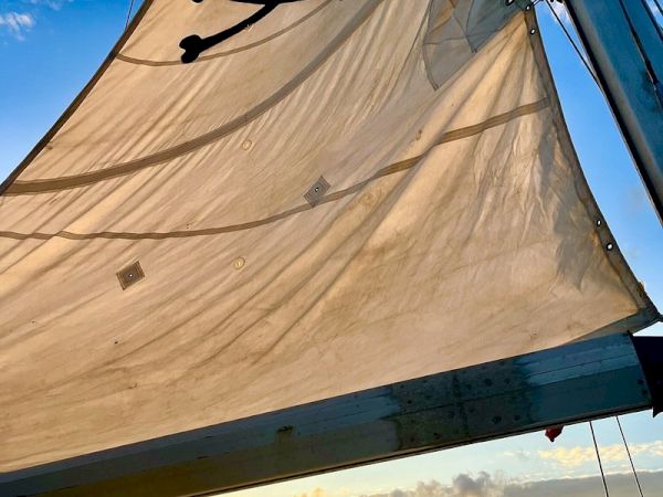 A sail with a skull and crossbones flag on a boat, sun setting over the horizon, blue sky with scattered clouds, rigging and sails catching wind.
