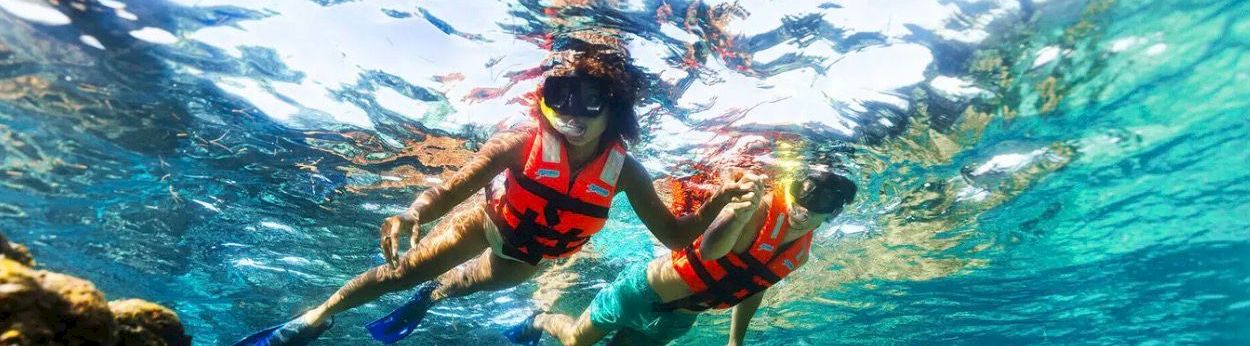Two people snorkeling underwater with red life jackets, fins, and clear blue water over a rocky seabed, exploring marine life.