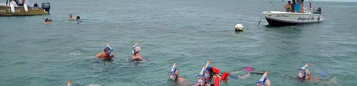 People snorkeling and swimming in clear ocean water near boats, with a cloudy sky and a buoy in the distance, colorful gear visible, everyone enjoying a sea excursion.