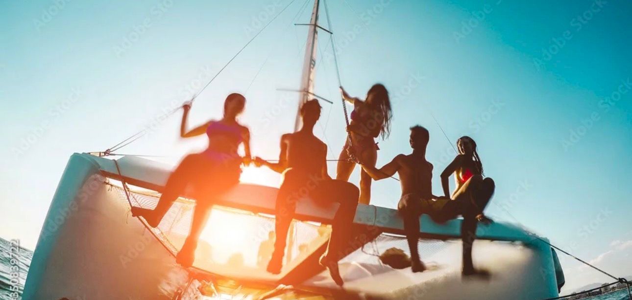A group of friends sits and stands on a small sailboat, sunlit, relaxing and enjoying the ocean at sunset.