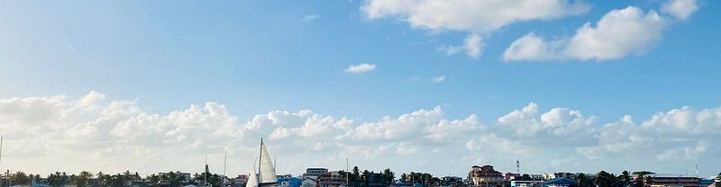 A tranquil harbor with small boats, a sailboat gliding by, and a distant shoreline under a bright blue sky with scattered clouds.