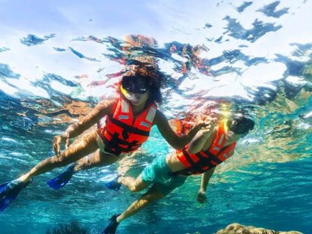 Two people snorkeling with orange life vests and fins, exploring a clear blue ocean over rocks and coral, sunlight sparkling above them.