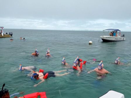 A group of people snorkeling and swimming in clear blue water near boats, with some wearing snorkeling gear and life vests, under a cloudy sky.