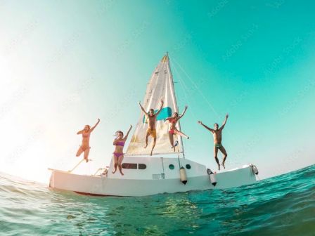 A group of friends jumps from a sailboat into clear turquoise water under a bright sky, enjoying a sunny seaside outing.