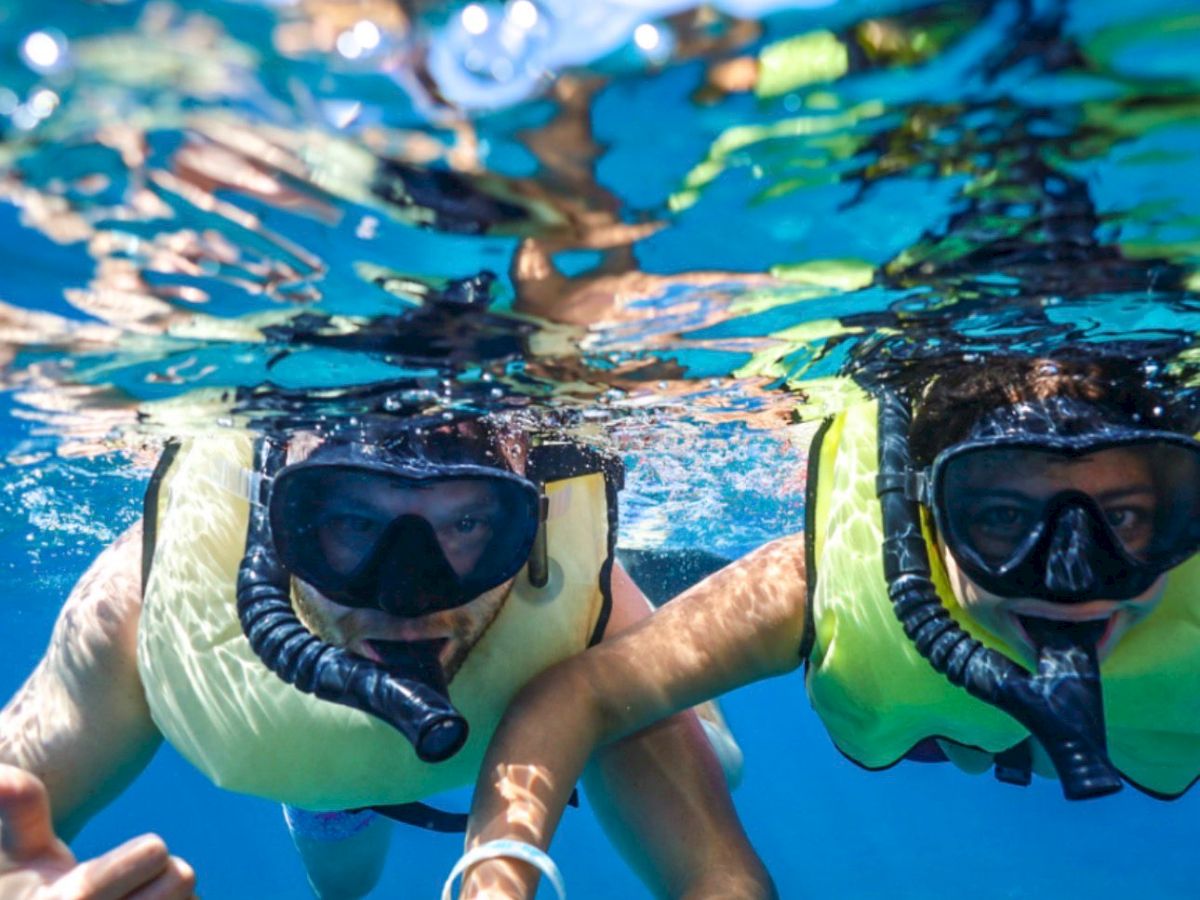 Two people snorkeling underwater, wearing life vests and masks, swimming toward the camera in clear blue water.
