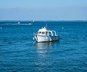A white motorboat cruises on a calm blue sea under a clear sky, with sailboats and small buoys visible in the distance.