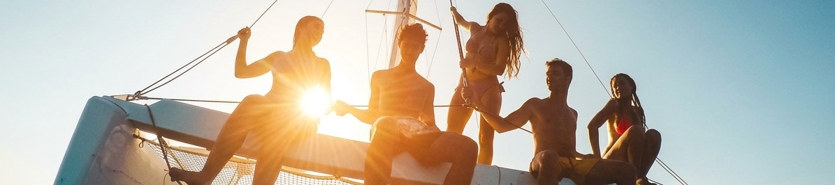 A group of friends relax on a catamaran deck, silhouetted by the setting sun over the ocean.