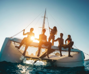 A group of friends relax on a catamaran deck, silhouetted by the setting sun over the ocean.