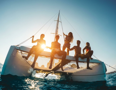 A group of friends relax on a catamaran deck, silhouetted by the setting sun over the ocean.