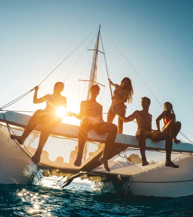 A group of friends relax on a catamaran deck, silhouetted by the setting sun over the ocean.