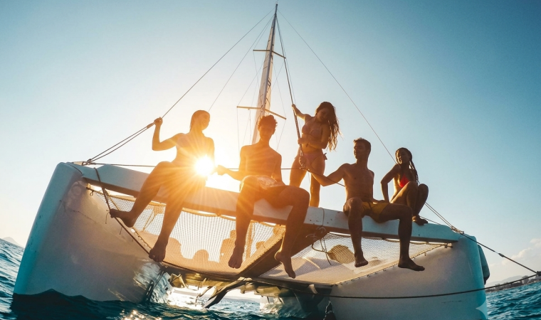 A group of friends relax on a catamaran deck, silhouetted by the setting sun over the ocean.