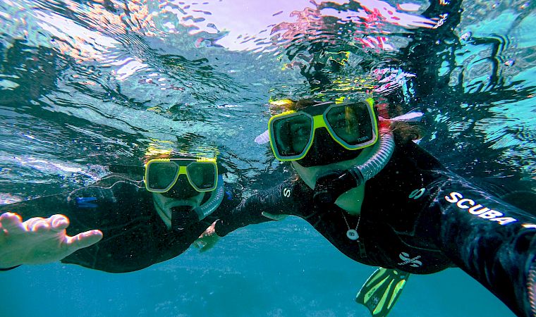 Two snorkelers swim underwater with green goggles and fins, pushing through bubbles and sunlight reflections.