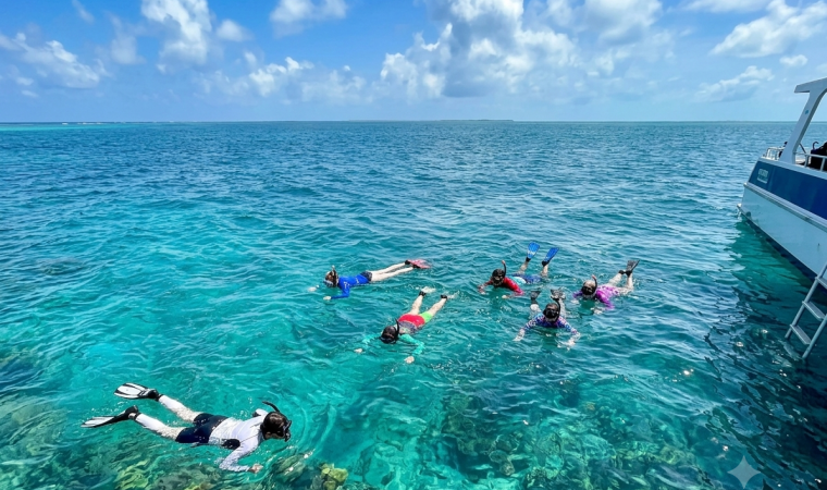 Snorkelers swim in clear turquoise ocean beside a boat under a bright blue sky dotted with clouds.